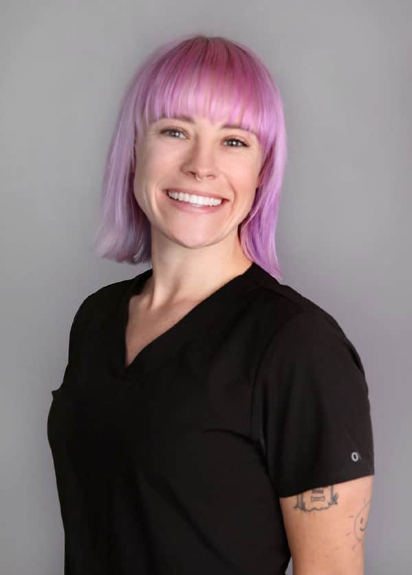 Person with short, straight pink hair and bangs, smiling. Wearing a black shirt that subtly complements their role as a compassionate veterinarian, while showing a tattoo on their left arm. Gray background.