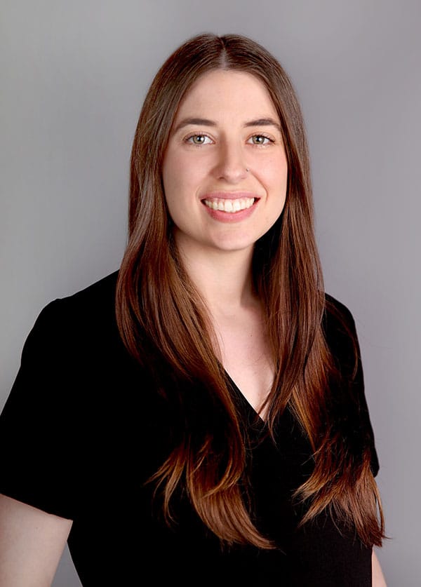 A woman with long brown hair and a nose ring smiles at the camera. As a veterinarian, she exudes warmth in her black V-neck top against a plain gray background.