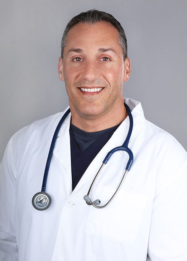 A smiling man with short, dark hair is wearing a white coat and a stethoscope around his neck, embodying the compassionate dedication of a skilled veterinarian. He stands confidently in front of a plain gray background.