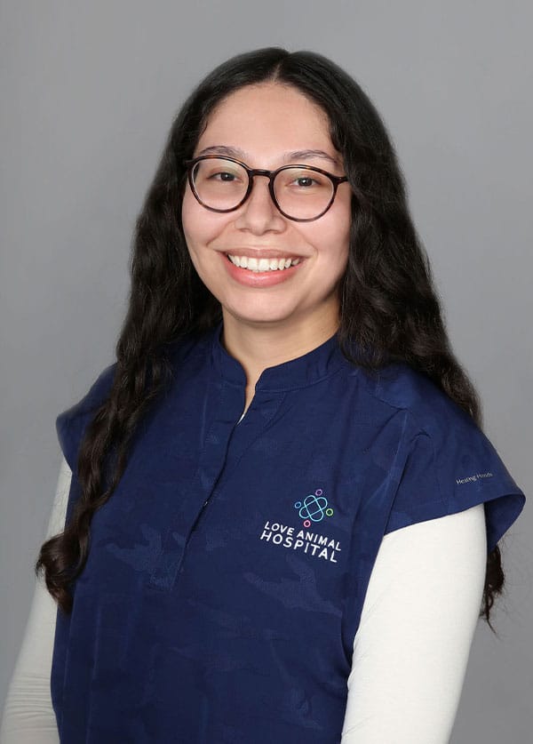 A veterinarian with long, dark curly hair and glasses smiles at the camera. They are wearing a dark blue shirt with a logo that reads "Love Animal Hospital." The background is a plain gray.